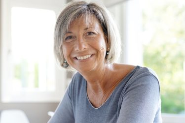 Ältere Frau mit grauem Haar lächelt, in grauem Oberteil vor unscharfem Fensterhintergrund.