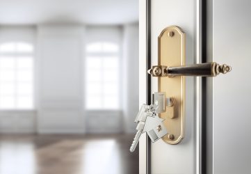Tür mit einem goldenen Türgriff und einem eingesteckten Schlüsselbund mit Hausanhänger. Im Hintergrund unscharfe Fenster.