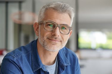 Ein Mann mit Brille und grauem Haar lächelt in die Kamera. Er trägt ein blaues Hemd.