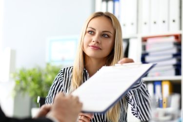 Frau in gestreifter Bluse hält ein Klemmbrett, im Hintergrund sind Bücherregale.
