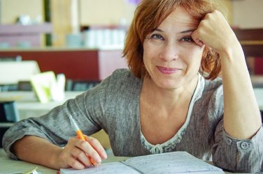 Eine Person sitzt mit einem Stift an einem Tisch und schaut lächelnd in die Kamera.