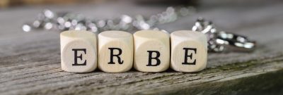 Holzwürfel mit Buchstaben "ERBE" auf einem Holztisch, unscharfe Kette im Hintergrund.