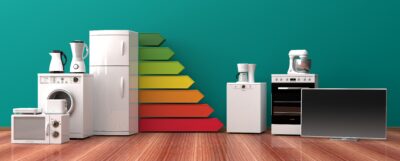 Haushaltsgeräte vor Energielabels in verschiedenen Farben. Holzfußboden, grüne Wand.