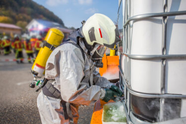 Person in Schutzanzug arbeitet an großem Behälter. Feuerwehr im Hintergrund unscharf.