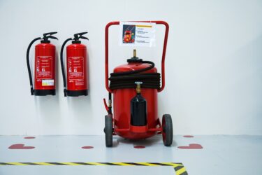 Zwei Wand-Feuerlöscher und ein mobiler Feuerlöscher auf Rädern in einem weißen Raum.