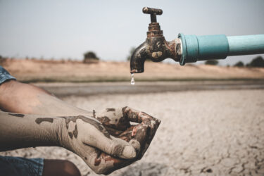 Eine Person hält Hände unter einen tropfenden Wasserhahn in einer ausgetrockneten Landschaft.