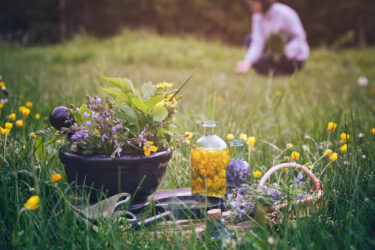 Bunte Blumen und Kräuter in einer Schale, Flaschen im Vordergrund, unscharfe Person sammelt im Hintergrund.