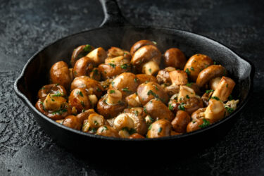 Gebratene Champignons mit Kräutern in einer schwarzen Pfanne auf dunklem Hintergrund.