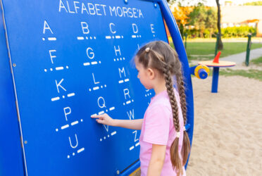 Kind zeigt auf Morsealphabet auf blauem Schild im Freien.