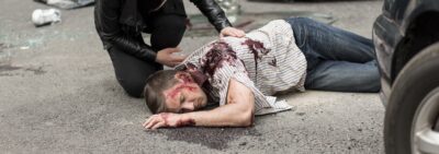 Eine verletzte Person liegt auf der Straße, während eine andere Person Hilfe leistet.