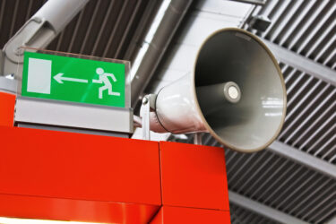 Grünes Notausgangsschild mit Pfeil und Figur neben großem Lautsprecher an roter Wand.