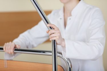 Person in weißem Kittel hält ein Geländer an einem Krankenbett.